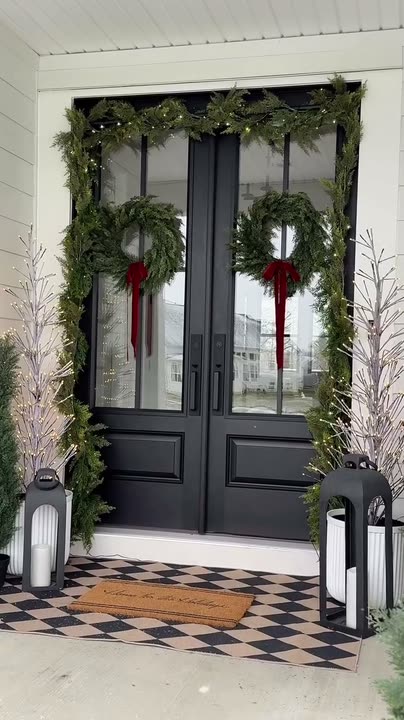 Reel by @modernfarmhouse_ohio: Black double front doors with glass panes, dressed with garland and red bows, on a white farmhouse porch.