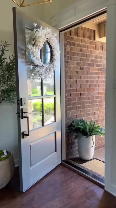 Reel by @modernhomedecor97: Grey-stained wood door with six glass panes and a white wreath, opened by hand from a brick entry.