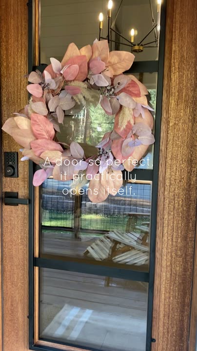 Reel by @surryplace: Natural mahogany front door with a full glass panel and a pink floral wreath, opening onto a covered porch. On-screen text: A door so perfect it practically opens itself.